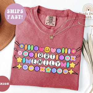 May include: Crimson-colored t-shirt with a beaded design featuring the name "EMILY" in white letters. The design includes colorful beads, hearts, flowers, and stars. A silver necklace with a pendant rests on the shirt. The words "SHIPS FAST!" are visible.
