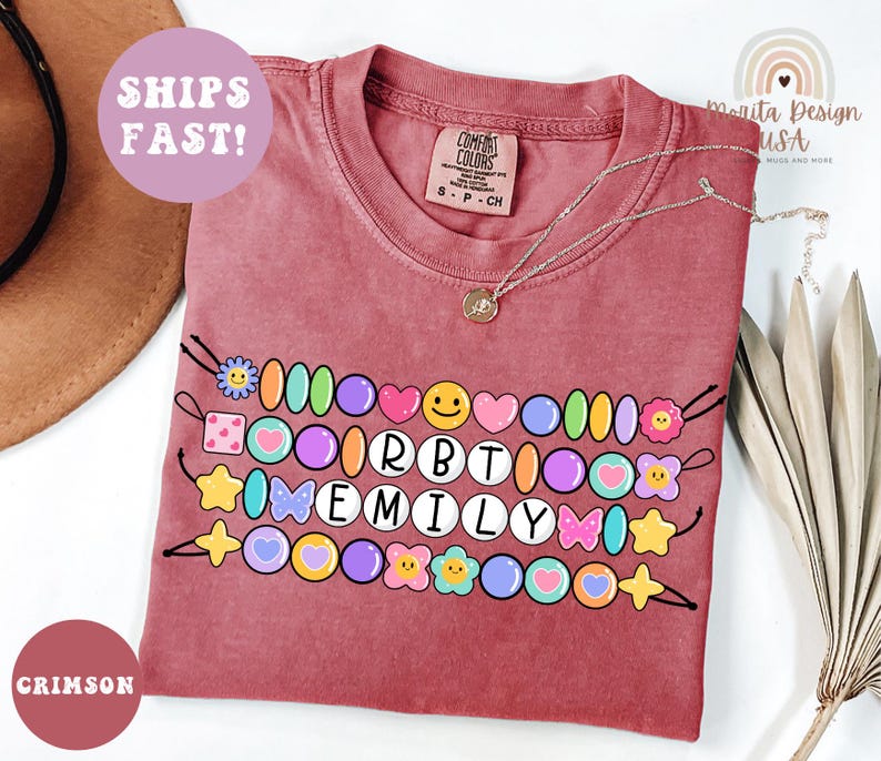 May include: Crimson-colored t-shirt with a beaded design featuring the name "EMILY" in white letters. The design includes colorful beads, hearts, flowers, and stars. A silver necklace with a pendant rests on the shirt. The words "SHIPS FAST!" are visible.