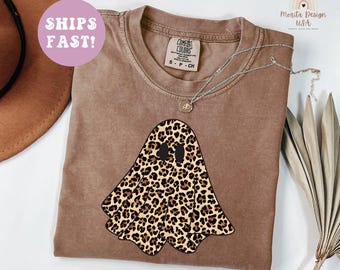 May include: A brown t-shirt featuring a leopard print ghost design. The shirt has a crew neck and short sleeves. The text "SHIPS FAST!" is in a pink circle in the upper left corner. A silver necklace with a pendant is on the shirt.