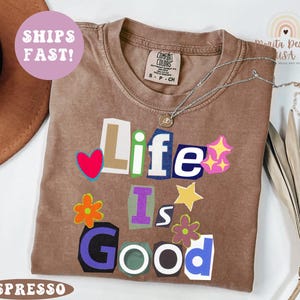 May include: Espresso-colored t-shirt with the phrase "Life Is Good" in a colorful, cut-out style. The shirt features a crew neck and short sleeves. A silver necklace with a pendant is draped on the shirt. The shirt is labeled "Comfort Colors".