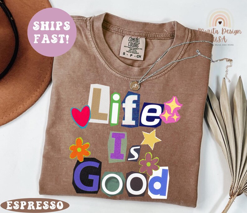 May include: Espresso-colored t-shirt with the phrase "Life Is Good" in a colorful, cut-out style. The shirt features a crew neck and short sleeves. A silver necklace with a pendant is draped on the shirt. The shirt is labeled "Comfort Colors".