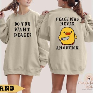 K&ouml;nnte beinhalten: Ein beiger Pullover mit dem Text "Do you want peace?" auf der Vorderseite und "Peace was never an option" mit einem Cartoon-gelben K&uuml;ken, das ein Messer h&auml;lt, auf der R&uuml;ckseite.