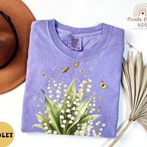 May include: A violet-colored Comfort Colors t-shirt featuring a floral design of lily of the valley and butterflies. The shirt has a crew neck and short sleeves. A gold necklace is draped across the shirt. The shirt is next to a brown hat and dried leaves.