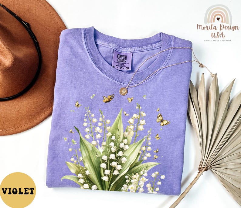 May include: A violet-colored Comfort Colors t-shirt featuring a floral design of lily of the valley and butterflies. The shirt has a crew neck and short sleeves. A gold necklace is draped across the shirt. The shirt is next to a brown hat and dried leaves.