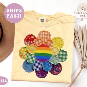 May include: A butter yellow t-shirt featuring a large flower graphic made of various fabric patches in plaid, floral, and gingham patterns. The center of the flower is a circular rainbow patch. The shirt has a Comfort Colors brand neck label.