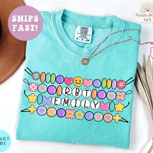 May include: A turquoise t-shirt with a beaded design spelling "EMILY" in white letters. The design includes colorful beads, hearts, stars, and flowers. A gold necklace rests on the shirt. The shirt has a "Comfort Colors" tag.
