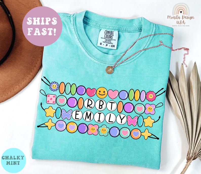 May include: A turquoise t-shirt with a beaded design spelling "EMILY" in white letters. The design includes colorful beads, hearts, stars, and flowers. A gold necklace rests on the shirt. The shirt has a "Comfort Colors" tag.