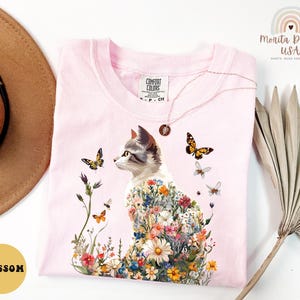 May include: Pink t-shirt featuring a floral cat design. The cat is surrounded by flowers and butterflies. The shirt has a tag that says "Comfort Colors".
