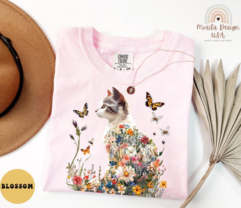 May include: Pink t-shirt featuring a floral cat design. The cat is surrounded by flowers and butterflies. The shirt has a tag that says "Comfort Colors".