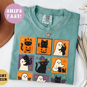 May include: Light green Comfort Colors t-shirt with a Halloween-themed graphic. The design features black cats, ghosts, and pumpkins in orange and purple squares. A silver necklace is draped across the shirt. The shirt is labeled "LIGHT GREEN" and "SHIPS FAST!"