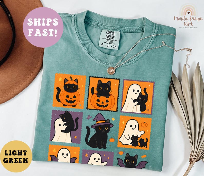 May include: Light green Comfort Colors t-shirt with a Halloween-themed graphic. The design features black cats, ghosts, and pumpkins in orange and purple squares. A silver necklace is draped across the shirt. The shirt is labeled "LIGHT GREEN" and "SHIPS FAST!"