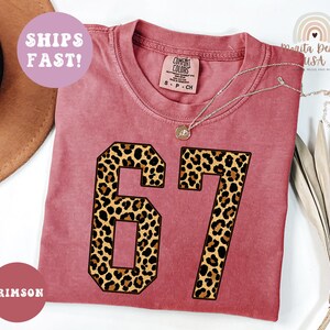 May include: Crimson-colored t-shirt with the number "67" in a leopard print design. The shirt has a crew neck and short sleeves. A silver necklace with a pendant is draped on the shirt. The words "SHIPS FAST!" are in a pink circle.