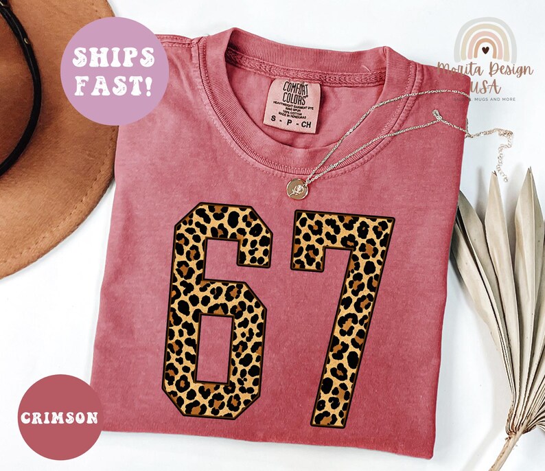 May include: Crimson-colored t-shirt with the number "67" in a leopard print design. The shirt has a crew neck and short sleeves. A silver necklace with a pendant is draped on the shirt. The words "SHIPS FAST!" are in a pink circle.