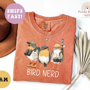 Op de afbeelding: Oranje-rood T-shirt met een afbeelding van drie vogelpersonages met hoeden, verrekijkers en camera's. De tekst "BIRD NERD" is in het wit gedrukt. Een gouden ketting is over het shirt gedrapeerd.