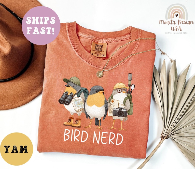 Op de afbeelding: Oranje-rood T-shirt met een afbeelding van drie vogelpersonages met hoeden, verrekijkers en camera's. De tekst "BIRD NERD" is in het wit gedrukt. Een gouden ketting is over het shirt gedrapeerd.