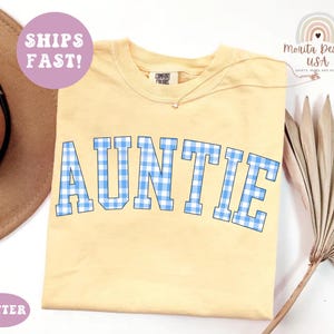 May include: A light yellow t-shirt with the word "AUNTIE" in blue and white gingham letters. The shirt has a crew neck and short sleeves. The text "Comfort Colors" is on the tag. A gold necklace with a star pendant is visible.