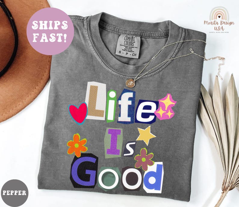 May include: A gray Comfort Colors t-shirt with the phrase "Life Is Good" in a colorful, cut-out style. The design includes a red heart, stars, and flowers. A silver necklace with a pendant is on the shirt.