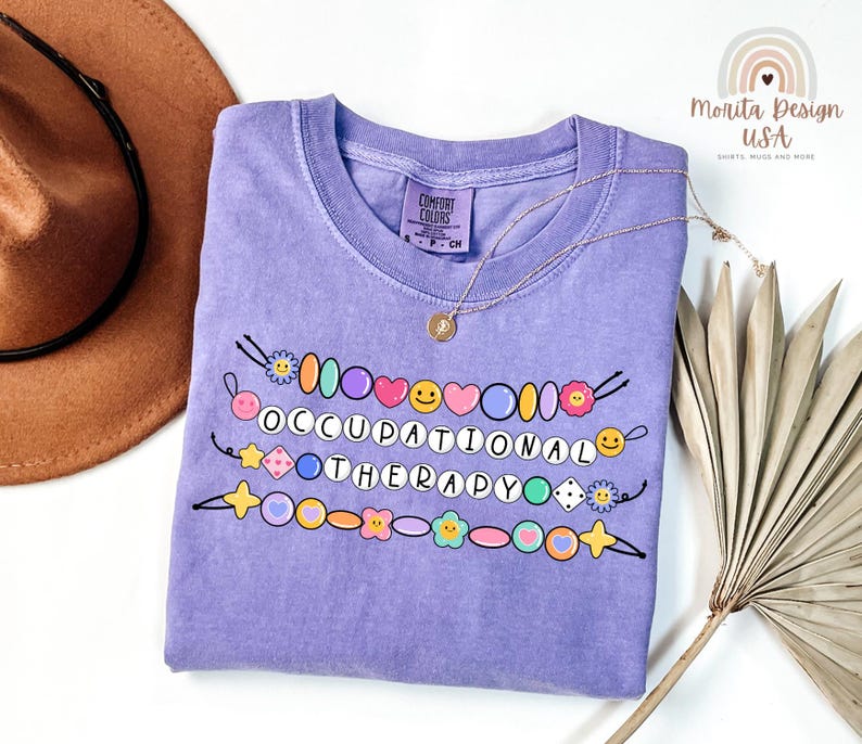 May include: A light purple t-shirt with the words "OCCUPATIONAL THERAPY" in white letters. The shirt features colorful beaded designs with smiley faces, hearts, and flowers. A gold necklace is draped across the shirt.