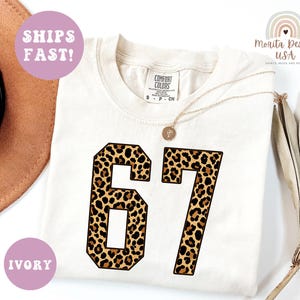 May include: Ivory-colored t-shirt with the number "67" in a leopard print design. The shirt has a crew neck and short sleeves. The text "SHIPS FAST!" and "IVORY" are in the image.