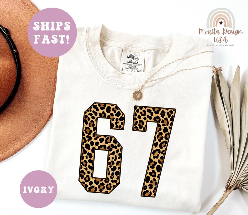 May include: Ivory-colored t-shirt with the number "67" in a leopard print design. The shirt has a crew neck and short sleeves. The text "SHIPS FAST!" and "IVORY" are in the image.