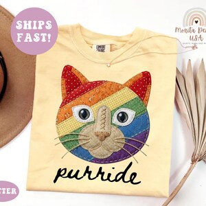 May include: A pale yellow t-shirt featuring a large, colorful cat face graphic made of rainbow-patterned fabric patches. The cat has white eyes with black pupils, a tan nose, and whiskers. Below the cat, the word purride is printed in black.