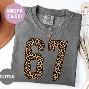 May include: A gray Comfort Colors t-shirt with the number "67" in a leopard print design. The shirt has a crew neck and short sleeves. The text "SHIPS FAST!" is in a pink circle. The shirt is labeled "PEPPER".