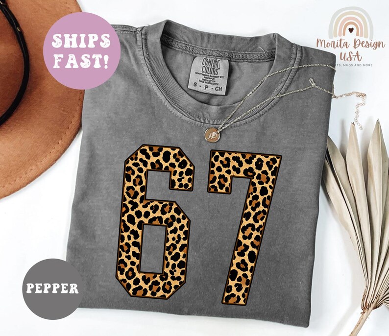 May include: A gray Comfort Colors t-shirt with the number "67" in a leopard print design. The shirt has a crew neck and short sleeves. The text "SHIPS FAST!" is in a pink circle. The shirt is labeled "PEPPER".