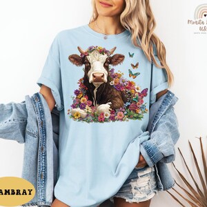 以下が含まれることがあります： 薄い青色のTシャツに、花冠をかぶった茶色と白の牛が花に囲まれているグラフィックがプリントされています。牛の周りを蝶が飛んでいます。画像の左下隅に「CHAMBRAY」というテキストが表示されています。