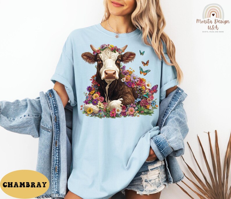 以下が含まれることがあります： 薄い青色のTシャツに、花冠をかぶった茶色と白の牛が花に囲まれているグラフィックがプリントされています。牛の周りを蝶が飛んでいます。画像の左下隅に「CHAMBRAY」というテキストが表示されています。