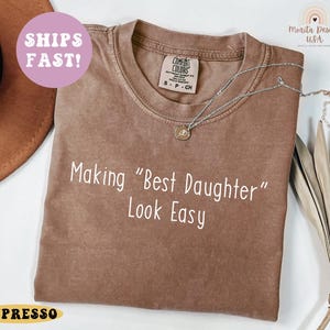 May include: A brown t-shirt with the text "Making "Best Daughter" Look Easy" in white. The shirt has a crew neck and short sleeves. A silver necklace with a pendant is draped on the shirt. The shirt has a vintage look.