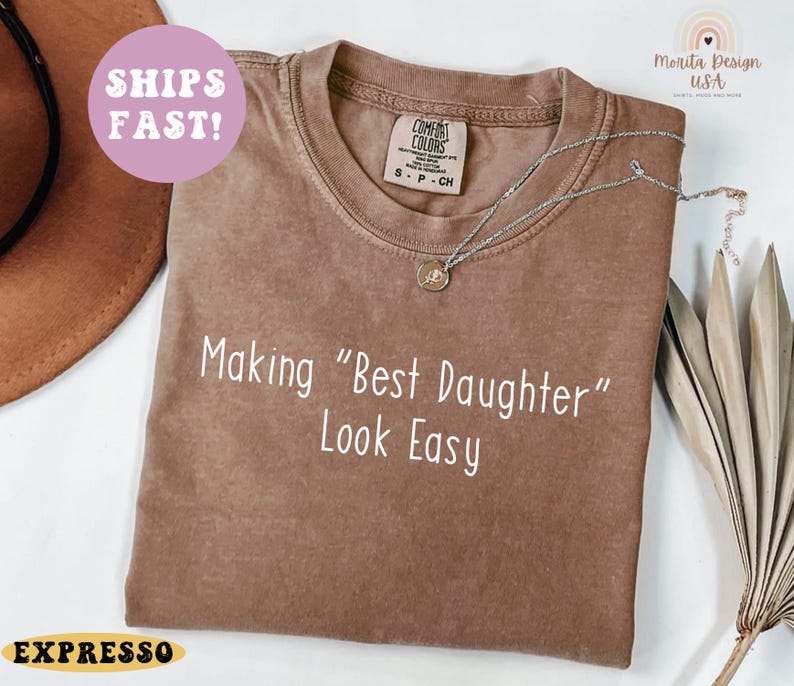 May include: A brown t-shirt with the text "Making "Best Daughter" Look Easy" in white. The shirt has a crew neck and short sleeves. A silver necklace with a pendant is draped on the shirt. The shirt has a vintage look.