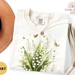 May include: Ivory-colored t-shirt with a floral design featuring white lily of the valley flowers and butterflies. The shirt has a crew neck and short sleeves. The brand name "Comfort Colors" is visible on the tag.