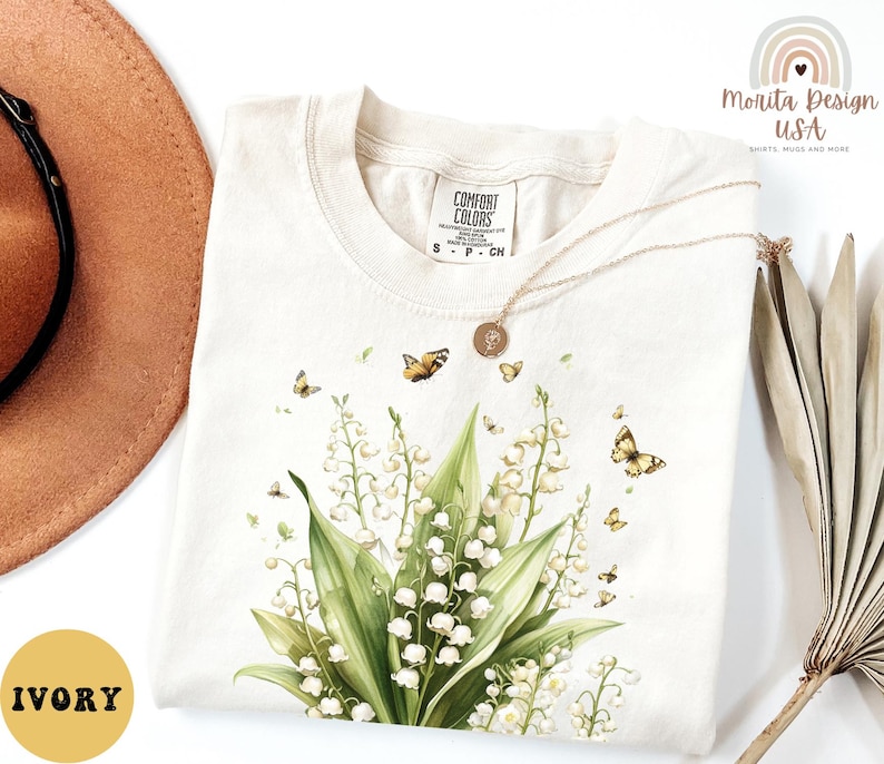 May include: Ivory-colored t-shirt with a floral design featuring white lily of the valley flowers and butterflies. The shirt has a crew neck and short sleeves. The brand name "Comfort Colors" is visible on the tag.