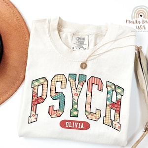 Peut inclure: T-shirt crème avec le mot "PSYCH" dans un motif patchwork, avec des motifs rouges, verts et bleus. En dessous du mot "PSYCH" se trouve le nom "OLIVIA" dans un ovale rouge. Un collier doré et un chapeau marron sont également visibles.