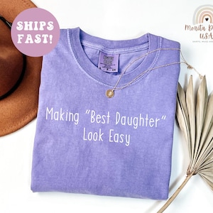 May include: A light purple Comfort Colors t-shirt with the phrase "Making "Best Daughter" Look Easy" in white. A gold necklace and a brown hat are also visible. The shirt has a crew neck and a small tag.
