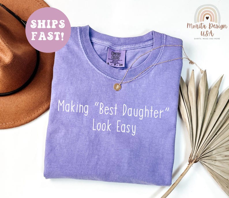 May include: A light purple Comfort Colors t-shirt with the phrase "Making "Best Daughter" Look Easy" in white. A gold necklace and a brown hat are also visible. The shirt has a crew neck and a small tag.