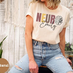 May include: A woman wearing a light beige t-shirt with the text "NEURO Emily" and a stylized brain graphic with floral accents. The shirt is a natural color.