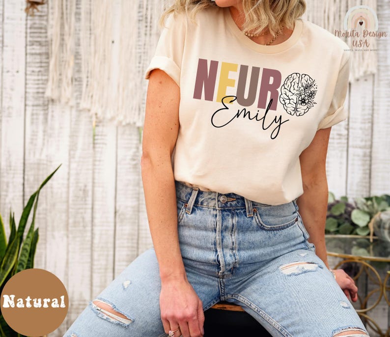 May include: A woman wearing a light beige t-shirt with the text "NEURO Emily" and a stylized brain graphic with floral accents. The shirt is a natural color.