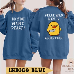 K&ouml;nnte beinhalten: Zwei indigo-blaue Sweatshirts mit wei&szlig;em Text. Ein Sweatshirt sagt "Willst du Frieden?" und das andere sagt "Frieden war nie eine Option." Beide Sweatshirts zeigen ein Cartoon-gelbes K&uuml;ken, das ein Messer h&auml;lt.