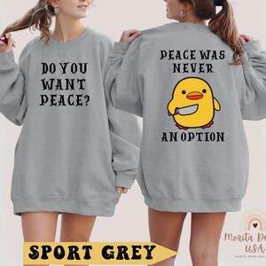 K&ouml;nnte beinhalten: Ein graues Sweatshirt mit dem Text "Do you want peace?" auf der Vorderseite und "Peace was never an option" mit einem Cartoon-K&uuml;ken, das ein Messer h&auml;lt, auf der R&uuml;ckseite. Das Sweatshirt ist in Sportgrau gehalten.