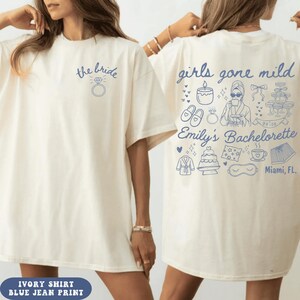 Puede incluir: Una camiseta color marfil con dibujos de l&iacute;neas azules. El frente muestra el texto "the bride" sobre un icono de anillo. La parte posterior presenta el texto "girls gone mild Emily's Bachelorette Miami, FL" rodeado de iconos de bata, pantuflas y pastel.
