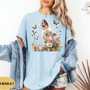 May include: A light blue t-shirt featuring a graphic of a white cat with flowers and butterflies. The cat is sitting on a bed of flowers. The text "CHAMBRAY" is visible in the bottom left corner of the image.