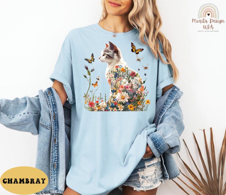 May include: A light blue t-shirt featuring a graphic of a white cat with flowers and butterflies. The cat is sitting on a bed of flowers. The text "CHAMBRAY" is visible in the bottom left corner of the image.