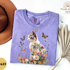 May include: A purple t-shirt featuring a floral cat design. The cat is surrounded by flowers and butterflies. The shirt has the text "Comfort Colors" on the tag.