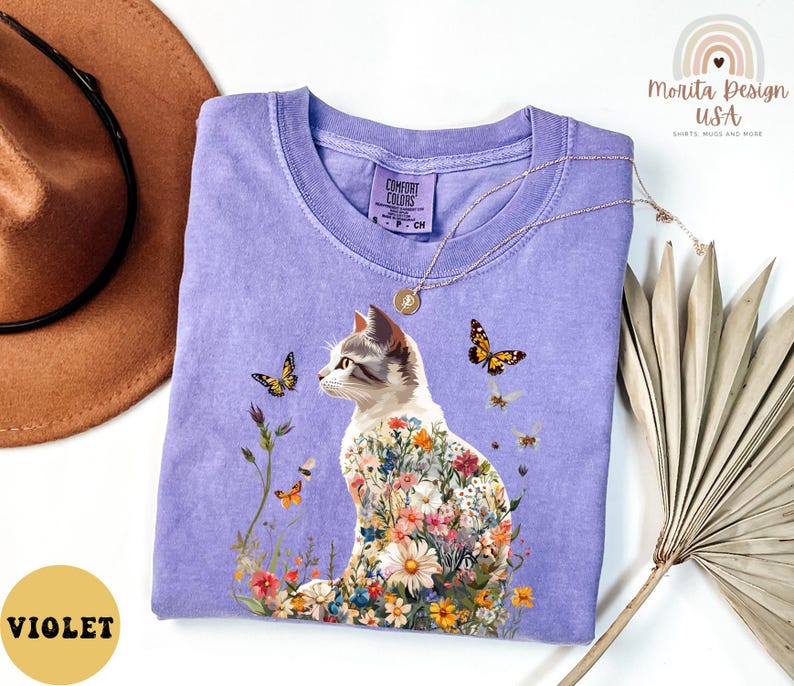 May include: A purple t-shirt featuring a floral cat design. The cat is surrounded by flowers and butterflies. The shirt has the text "Comfort Colors" on the tag.