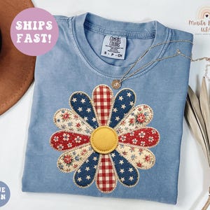May include: A light blue Comfort Colors t-shirt with a large daisy applique. The daisy features red, white, and blue patterned petals and a yellow center. A gold necklace rests on the shirt. The text "SHIPS FAST!" is in a pink circle.