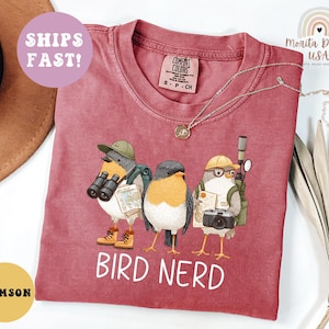 Op de afbeelding: Karmizijnrood T-shirt met een grillige illustratie van drie vogelpersonages uitgerust met verrekijkers, camera's en kaarten. De tekst "BIRD NERD" is in het wit gedrukt. Het shirt heeft een Comfort Colors label.