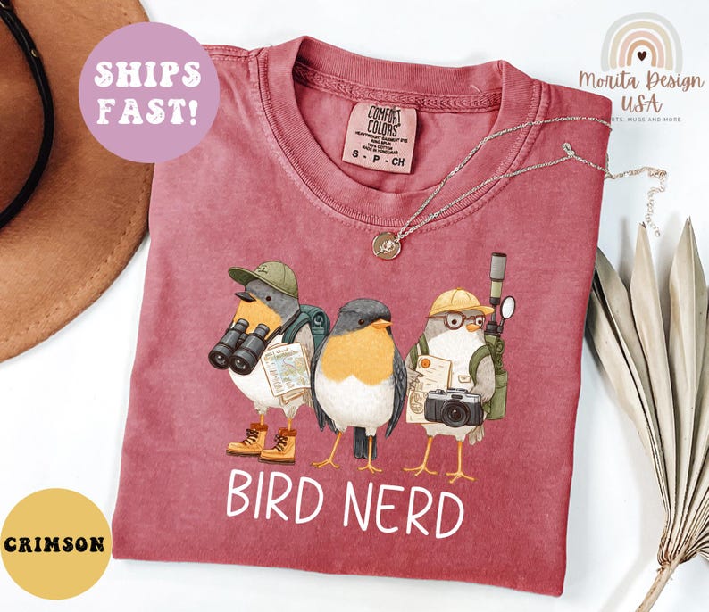Op de afbeelding: Karmizijnrood T-shirt met een grillige illustratie van drie vogelpersonages uitgerust met verrekijkers, camera's en kaarten. De tekst "BIRD NERD" is in het wit gedrukt. Het shirt heeft een Comfort Colors label.