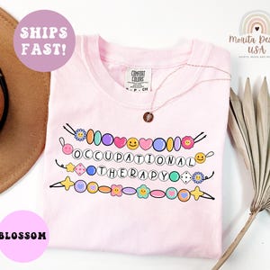 May include: A light pink t-shirt with the words "OCCUPATIONAL THERAPY" in a colorful beaded design. The shirt also has the text "SHIPS FAST!" and "BLOSSOM" in a pink circle.