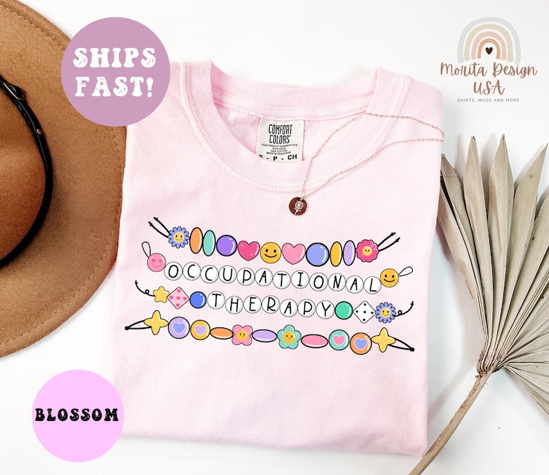 May include: A light pink t-shirt with the words "OCCUPATIONAL THERAPY" in a colorful beaded design. The shirt also has the text "SHIPS FAST!" and "BLOSSOM" in a pink circle.